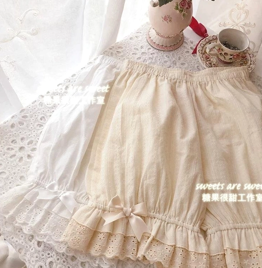Overhead flat lay of beige and white premium lolita cotton bloomers with multi-layer ruffled embroidered lace cuffs, decorative bow accents, vertical stripe fabric pattern texture, styled on vintage delicate lace tablecloth with antique tea service and romantic period aesthetic