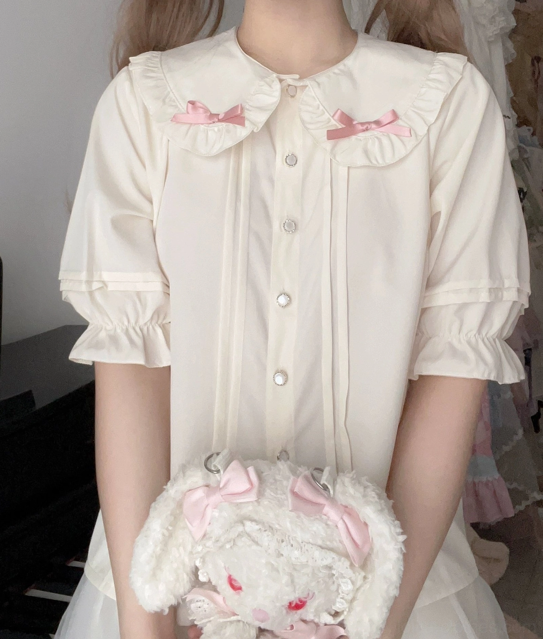 Close-up of cream peter pan collar blouse with pink bow details, puffed short sleeves, front button placket, paired with plush crossbody bag