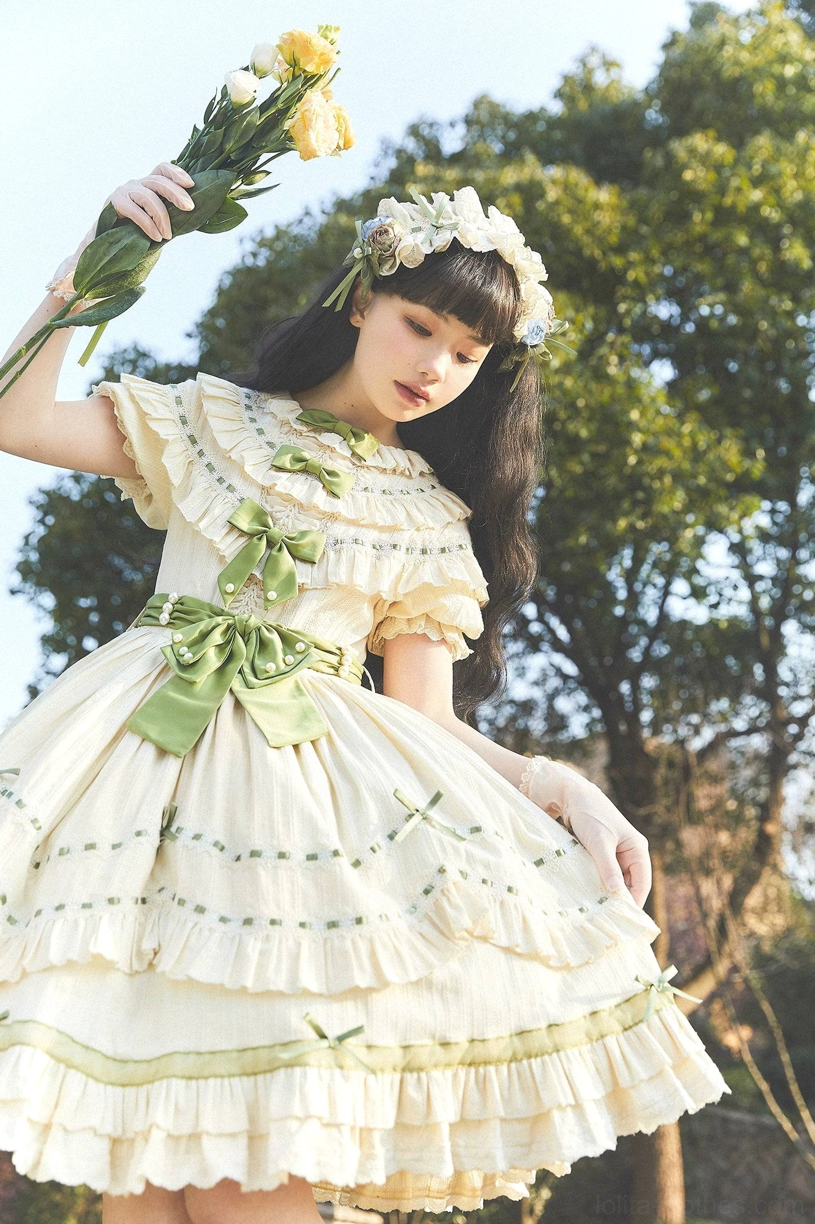 Close-up three-quarter front view of country lolita dress showing ruffled Peter Pan collar, large sage green waist bow, and tiered ruffle skirt with embroidery details and flower crown
