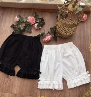 Overhead flat lay of elegant lolita elastic waist cotton bloomers featuring black and white variants with multi-layer ruffled embroidered lace hems, displayed on vintage wooden table with pink roses and decorative ornamental birdcage, showcasing premium cotton craftsmanship and color palette options