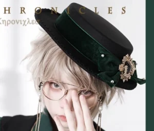 Lolita flat bonnet in forest green velvet worn by blonde male model with round glasses, showing gold brooch and pearl embellishments, close-up half-body portrait
