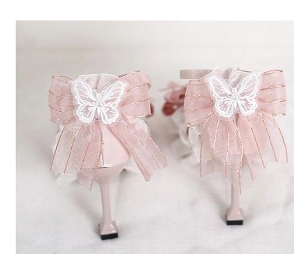 Close-up detail of lolita shoe bow decoration featuring layered sheer pink ribbon, white embroidered butterfly center, gold hardware accents, and delicate ruffled mesh trim on plain light background