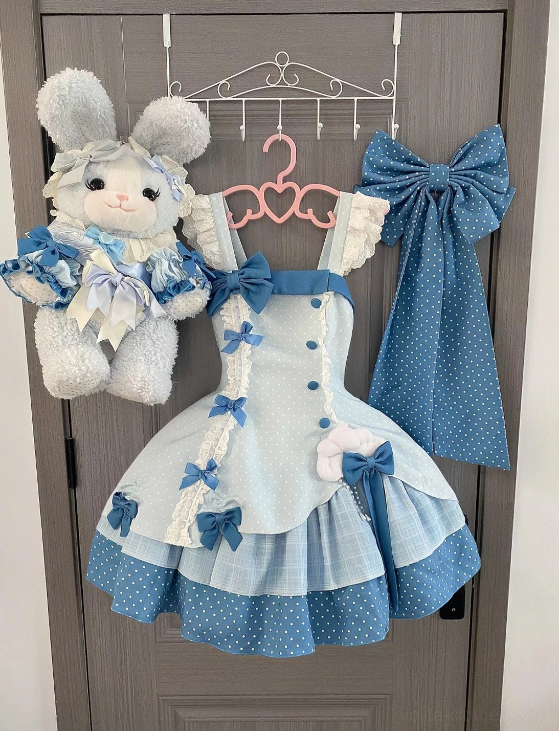 Lolita JSK jumper skirt dress hanging display with matching accessories and plush rabbit doll wearing coordinating mini dress, showcasing polka dot blue bows, ruffled shoulder straps, tiered ruffled hem details
