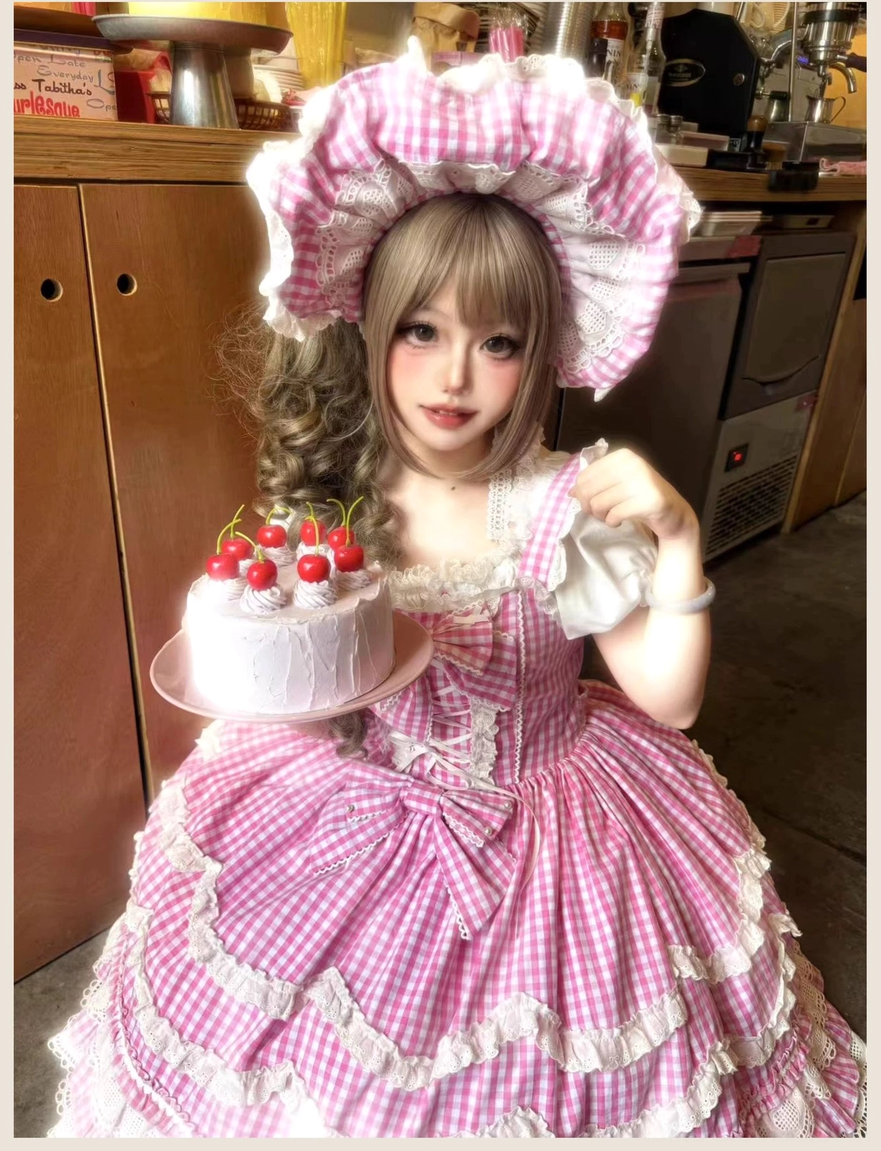 Pink gingham old school Lolita JSK dress with matching large bonnet and corset lace-up front, close-up full-body front view in cozy cafe kitchen setting with cherry cake dessert plate styling