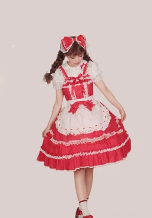 Red and white classic Lolita JSK dress on model with solid pink background, featuring tiered ruffles, apron-style panel, and coordinating red bow hair accessory