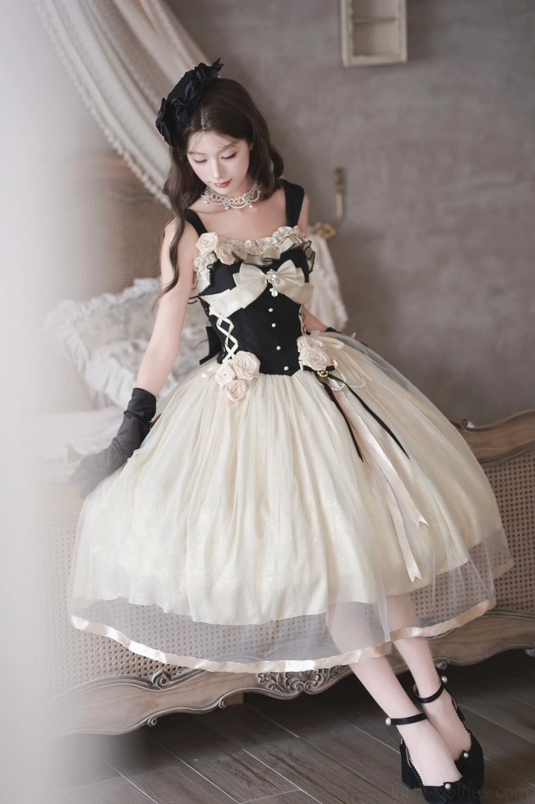 Side sitting full-body view of Classic Lolita corset jumper skirt on vintage bed, flowy tulle skirt with satin hem trim, corset bodice with bow and rose decorations, black gloves and heels