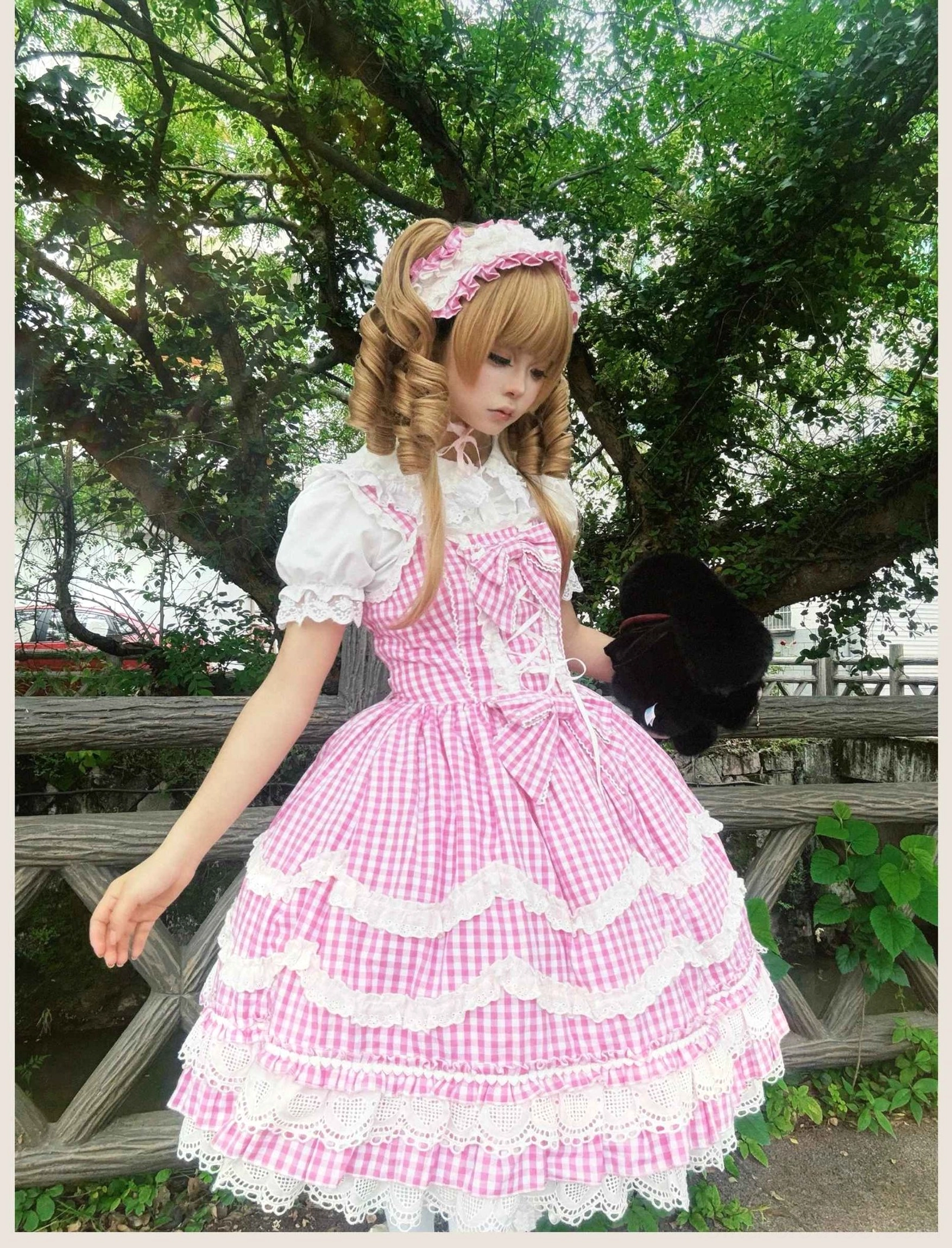 Pink gingham Lolita JSK dress featuring adjustable lace-up bodice and tiered ruffled skirt, outdoor full-body side view garden fence background with matching bonnet and black stuffed animal