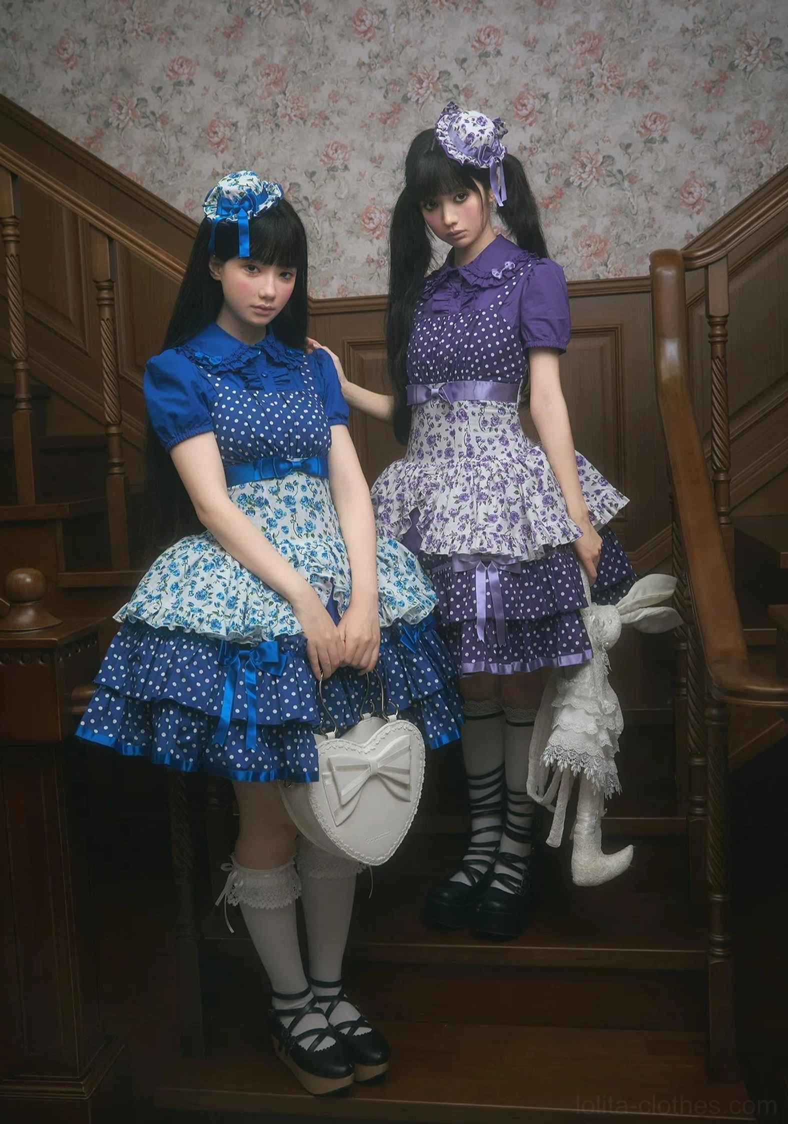 Blue and purple old school Lolita jumper skirts with Peter Pan collar blouses on wooden staircase, full-body side group view with two models wearing matching hair bows, knee-high socks, and Mary Jane loafers