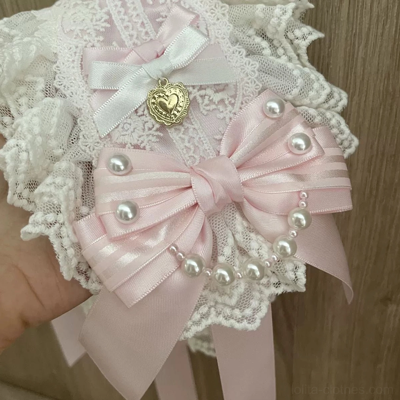 Detailed close-up of Lolita hairband bow design, large pink satin bow with small white satin bow, gold heart pendant, faux pearl embellishments on white lace base, flat lay
