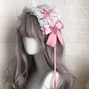 Delicate lolita lace headdress with dusty pink satin bows and long ribbon tails, worn by mannequin with ash purple wig against white brick wall, showcasing romantic ribbon bow design from front angle