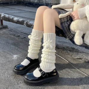 Off-white knitted lolita leg warmers with fuzzy pom-pom accents and ruched texture worn seated on outdoor rooftop with plaid skirt and black Mary Jane shoes