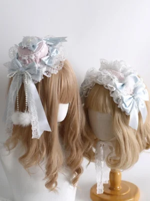 Lolita mini top hat KC and hair clip set with pom-pom tassel charms displayed on two mannequins with blonde wavy wigs, showing pink-white layered lace base with sky blue bows from side group perspective