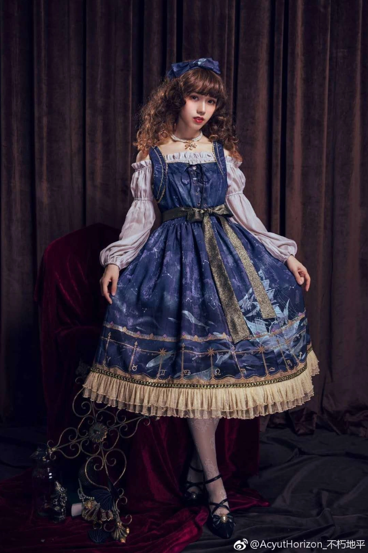 Lolita fashion model wearing royal blue dress with off-shoulder puff sleeves and ruffled hem coordinated with pearl necklace, full-body standing portrait in studio with dark red velvet backdrop, vintage royal lolita style