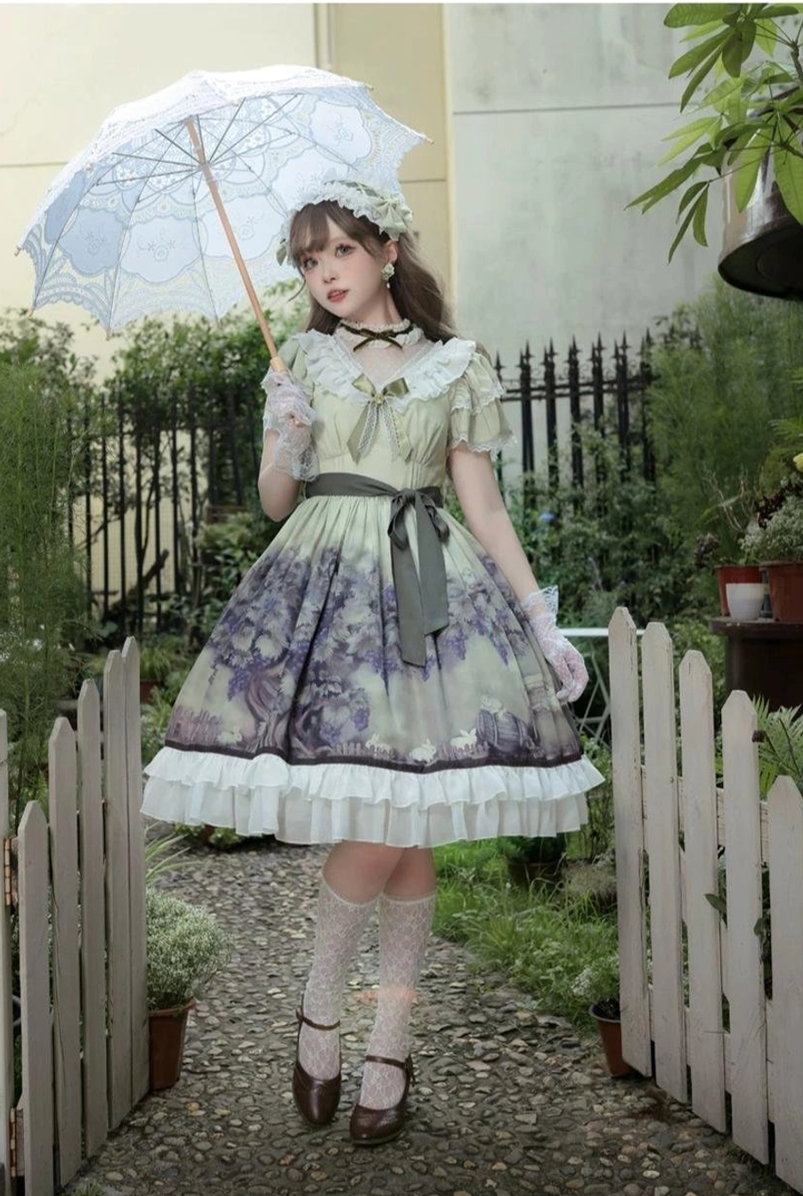 Front full walking view of sage green lolita OP dress on model strolling garden path with white picket fence background, wearing consistent grape floral print throughout dress, lace stockings and brown shoes