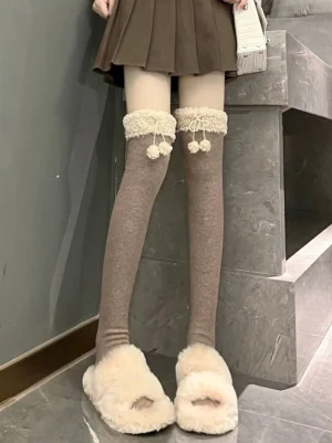 Kawaii winter lolita over-knee socks with taupe brown fuzzy lambswool trim and pom-pom cuff bow, worn over-the-knee with brown pleated skirt and fuzzy slippers on marble floor, front low-angle view of lolita fashion hosiery