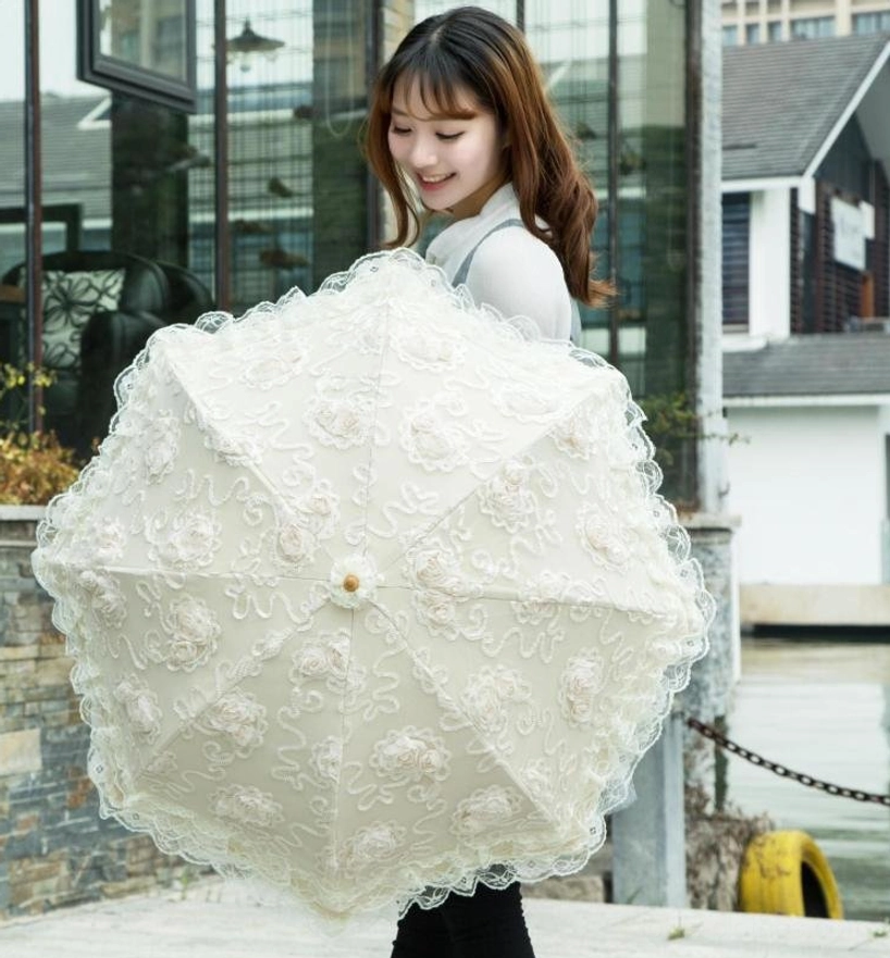 Front eye-level view of female model holding unfolded lolita lace parasol outdoors by waterside building, showcasing full coverage of 3D floral lace appliques, central wooden finial, cream white color