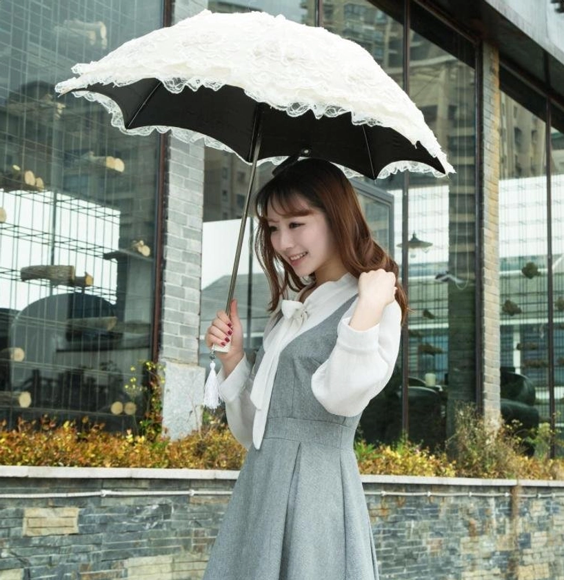 Female model holding unfolded lace parasol overhead outdoors against modern glass building background, showing white lace exterior with black sun-protective inner canopy and ruffled lace trim