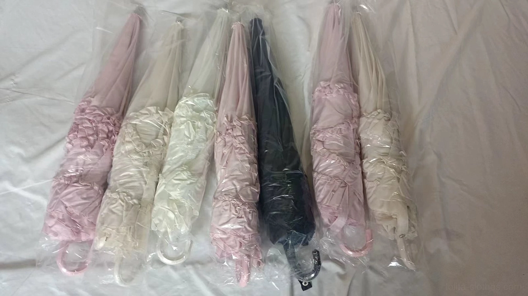 Ruffled Lolita parasols in retail packaging with pastel pink, beige, and black color options wrapped in protective plastic bags, overhead flat lay on white fabric
