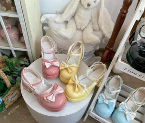 Close-up tabletop display of gradient Sweet Lolita platform heels with glossy patent leather vamp bow and heart-shaped block heel cutout, arranged on white stand with cute teddy bear and cozy bedroom decor background