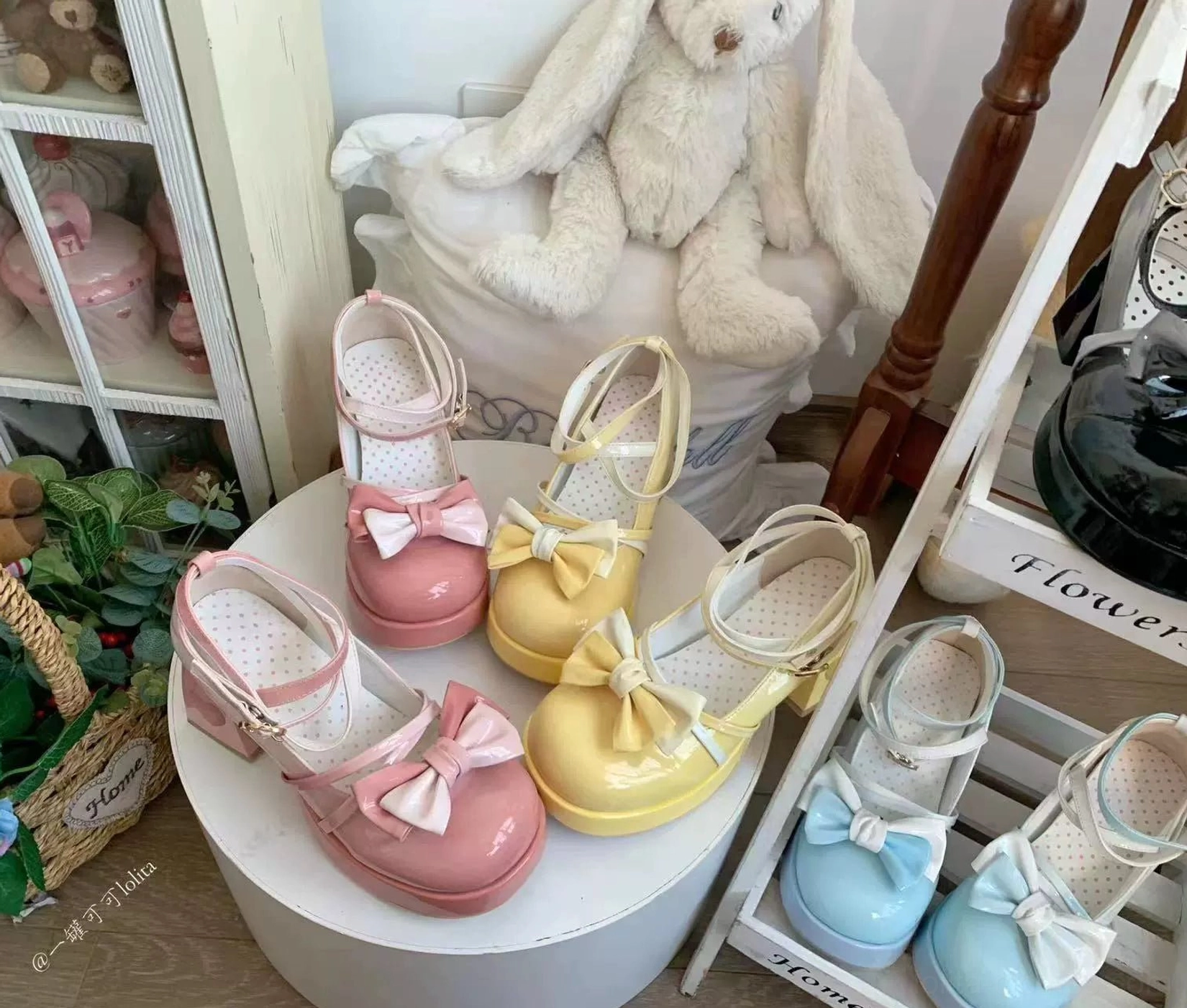 Close-up tabletop display of gradient Sweet Lolita platform heels with glossy patent leather vamp bow and heart-shaped block heel cutout, arranged on white stand with cute teddy bear and cozy bedroom decor background