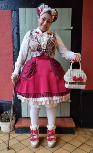 Female model wearing complete wine red tiered Lolita coordinate featuring voluminous petticoat base, matching tiered skirt with ruffled trim, patterned waistcoat and hair accessories, classic Lolita fashion styling