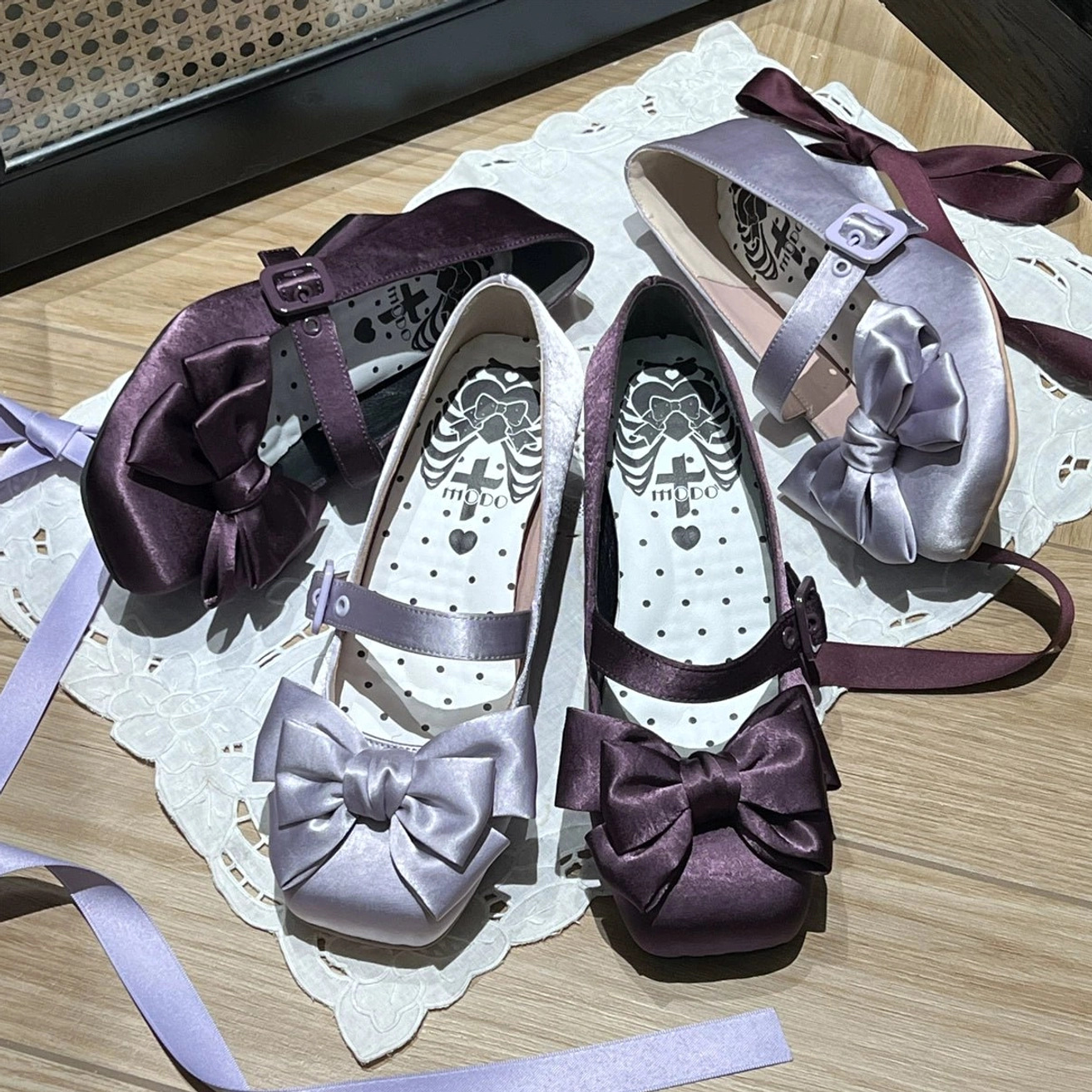 Overhead flat lay of three purple-toned lolita satin Mary Jane shoes with satin bow decorations and adjustable buckle ankle straps on lace placemat