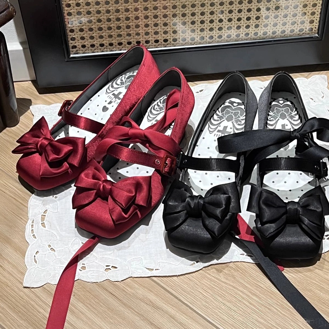 Top-down flat lay of burgundy and black lolita satin square toe shoes with oversized satin bows and ribbon ankle straps on wooden table with lace doily