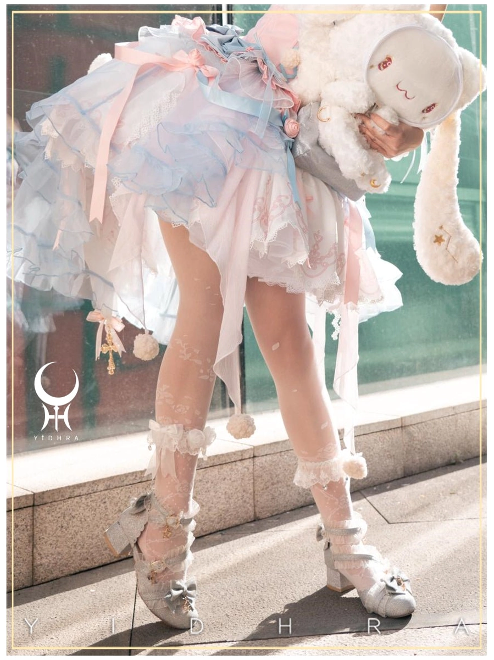 Low angle outdoor side shot of beige Lolita thin sheer tights with white lace ruffled ankle trims, modeled with pink-blue gradient sweet Lolita dress, silver Mary Jane shoes, and plush rabbit companion accessory