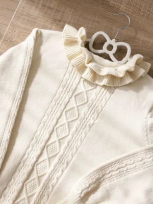 Close-up detail of ivory lolita sweater neckline featuring layered ruffled stand collar with front cable knit pattern, overhead macro photography showcasing textile quality and ruffle construction