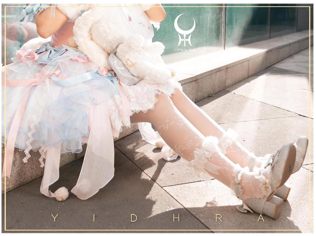 Low angle side shot of seated model wearing beige Lolita thin sheer tights with white lace ruffled ankle cuffs, pink-blue gradient sweet Lolita dress, beige block heel shoes, holding plush rabbit doll