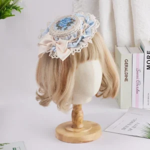 Delicate Lolita miniature hat featuring two-tone beige and light blue satin bow with blue floral embroidery medallion and white lace trim, displayed on styled mannequin head