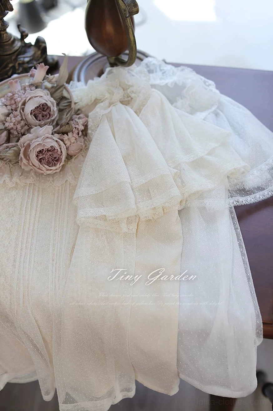 Flat lay close-up detail of classic lolita tulle blouse displaying cream beige polka-dot tulle texture, delicate ruffled flutter sleeves, lace trim elements, vertical pintuck pleats and vintage floral brooch accent