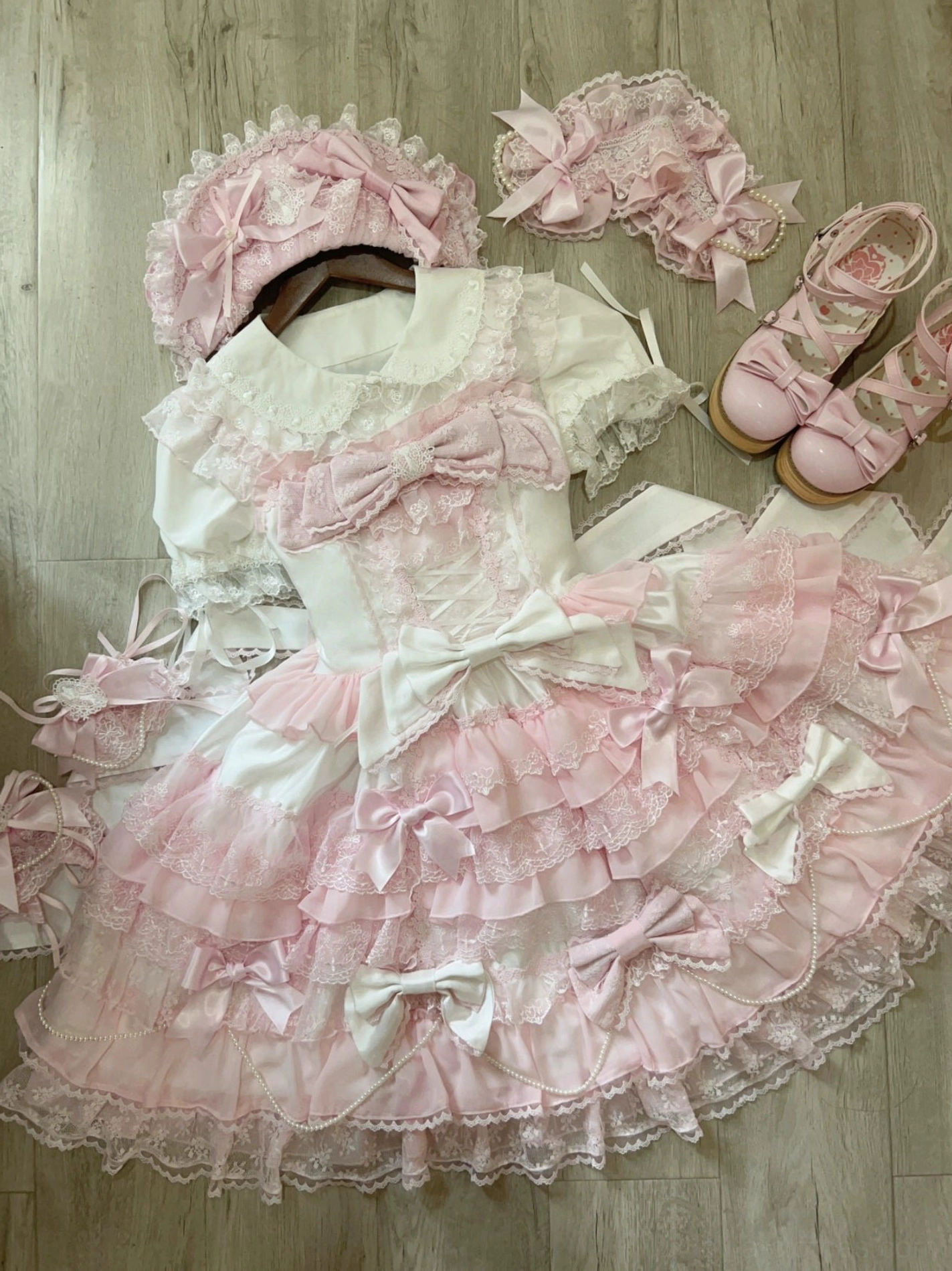Overhead flat lay of complete lolita outfit on wooden floor including dress bonnet hairband pink platform shoes and lace hem trim details