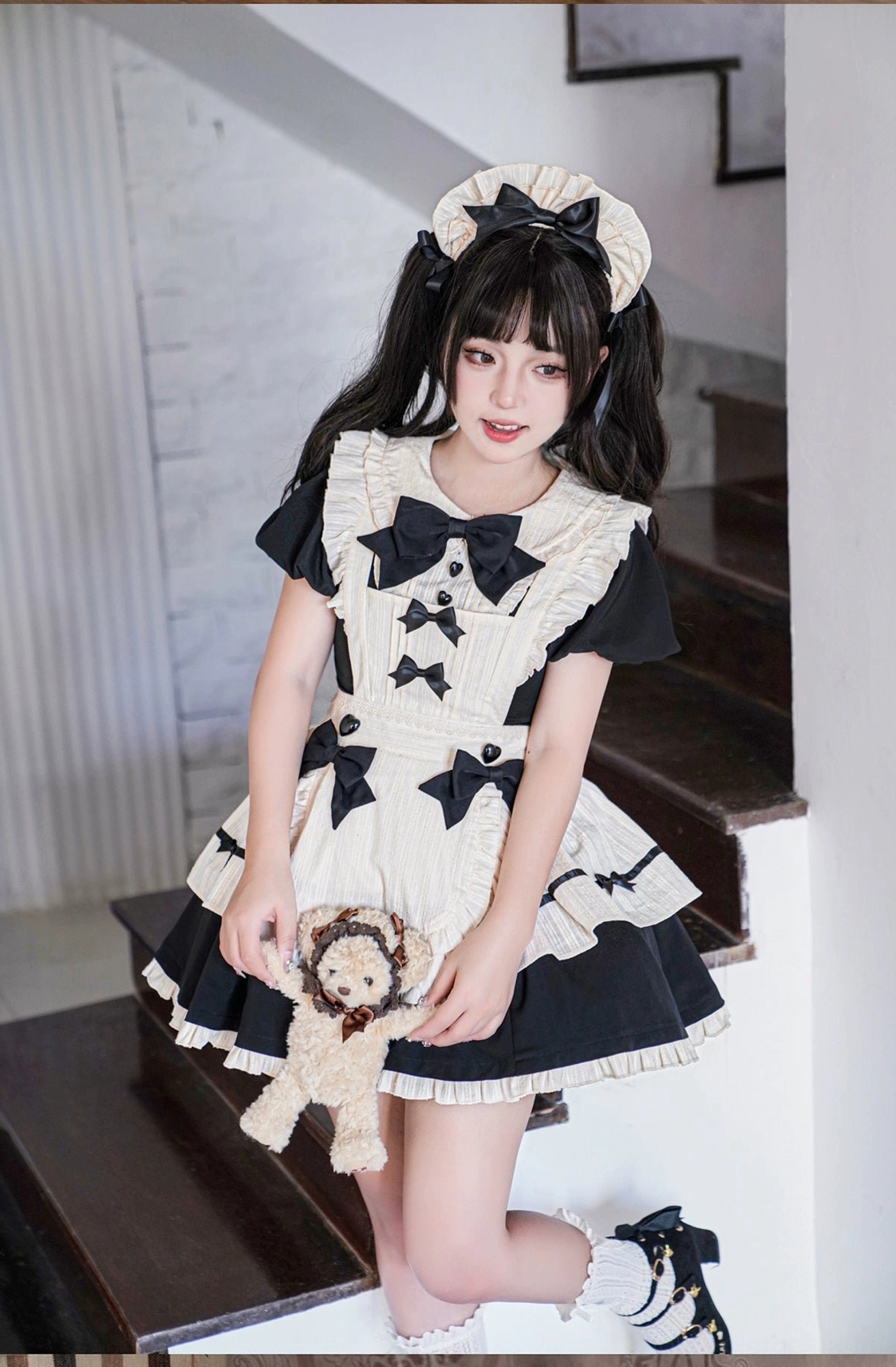 Low-angle full-body view of maid lolita apron dress on model holding plush toy showing multi-bow bodice and ruffled hem details kneeling on stairs