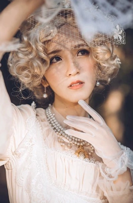 Curly vintage-style model in ivory ruffled lolita dress with white gloves wearing Manor Ball II pearl necklace and matching pearl earrings warm indoor setting