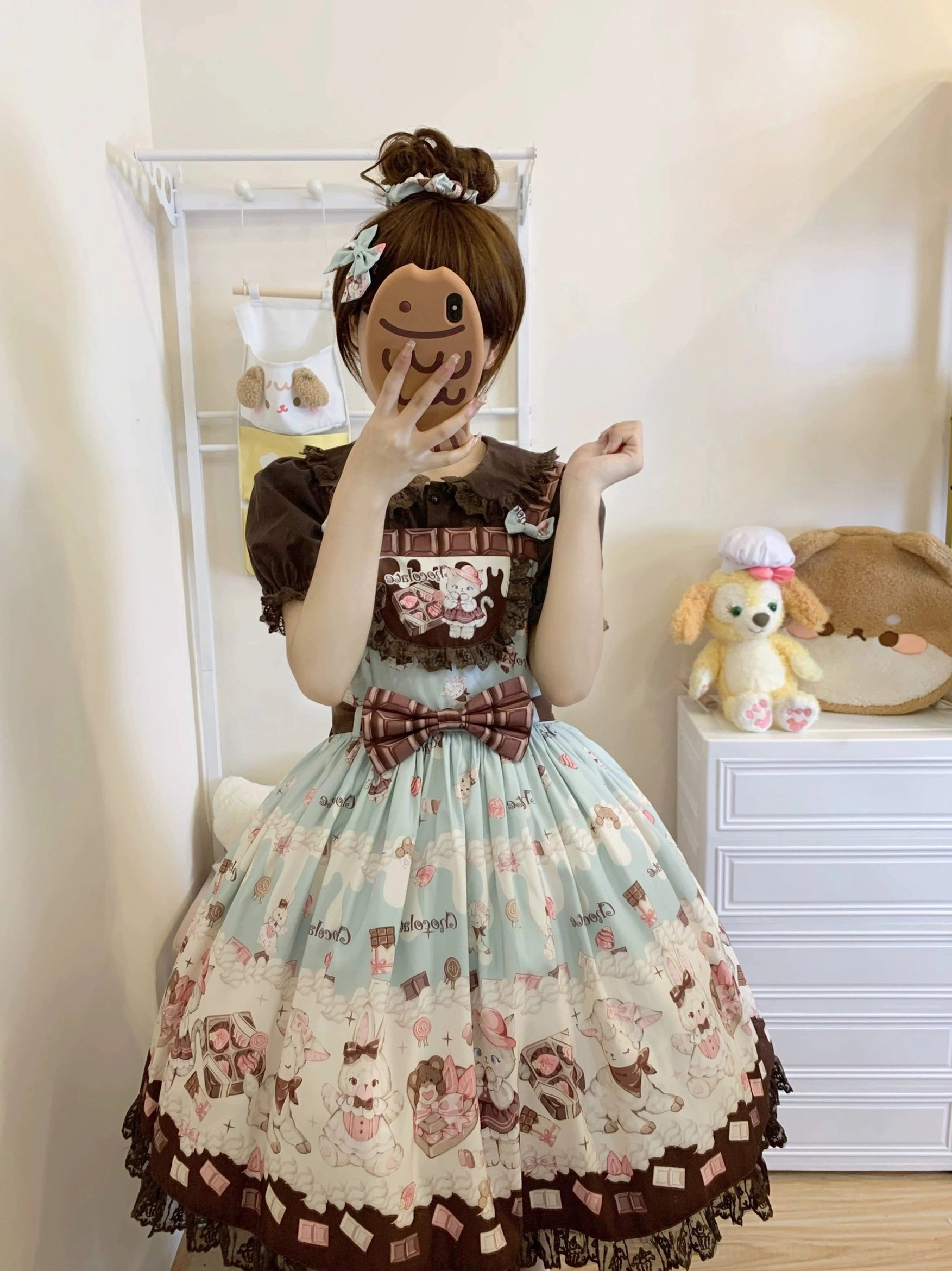 Model with topknot hairstyle wearing mint green and brown cat print Lolita salopette dress with waist bow detail and checkered printed skirt hem, half-body hidden face shot against white storage shelf backdrop