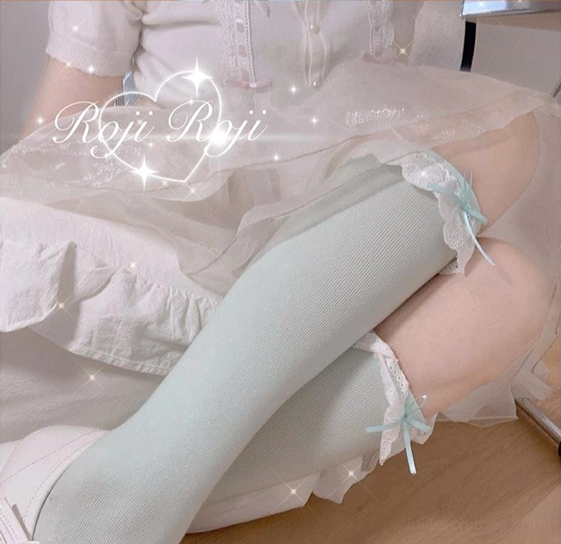 Mint green lace-trimmed sweet lolita stretch knit socks with tiny blue bow accents on lace cuff, close-up side lying view on ruffled bedding with white frilly blouse