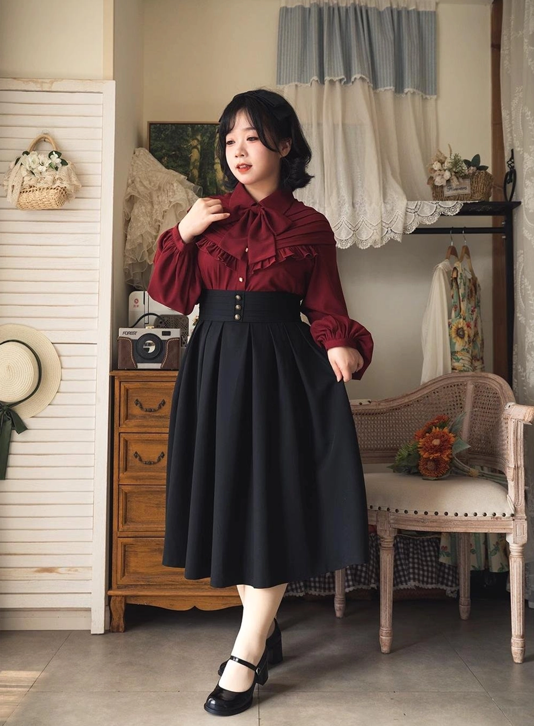 Model in three-quarter front view wearing vintage Lolita outfit with burgundy bow blouse, adjusting collar to show ruffled neck details and silhouette of high-waisted skirt against retro studio background