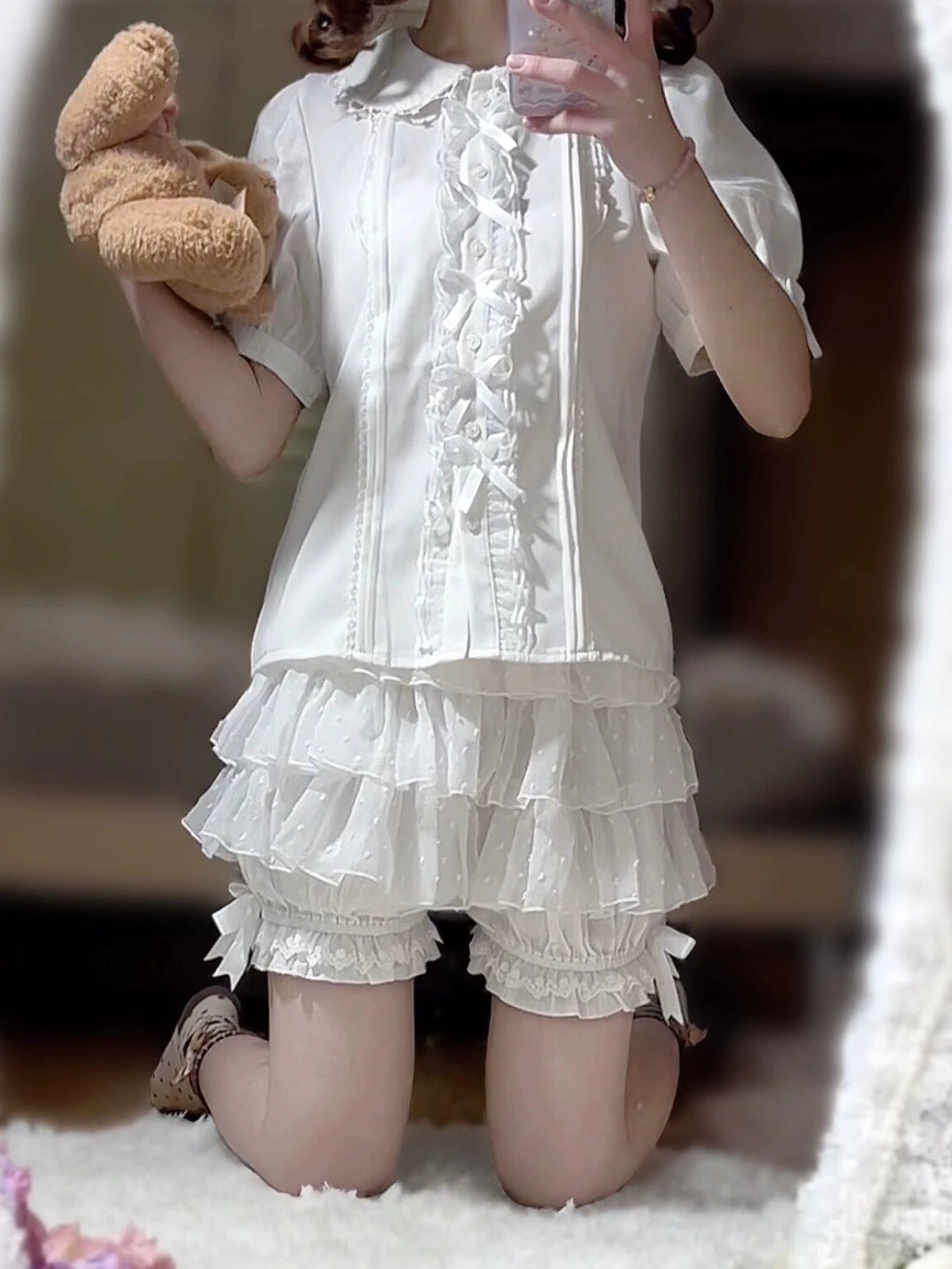 Model wearing white lolita bloomers styled with puff-sleeve blouse in front mirror selfie pose holding teddy bear indoors