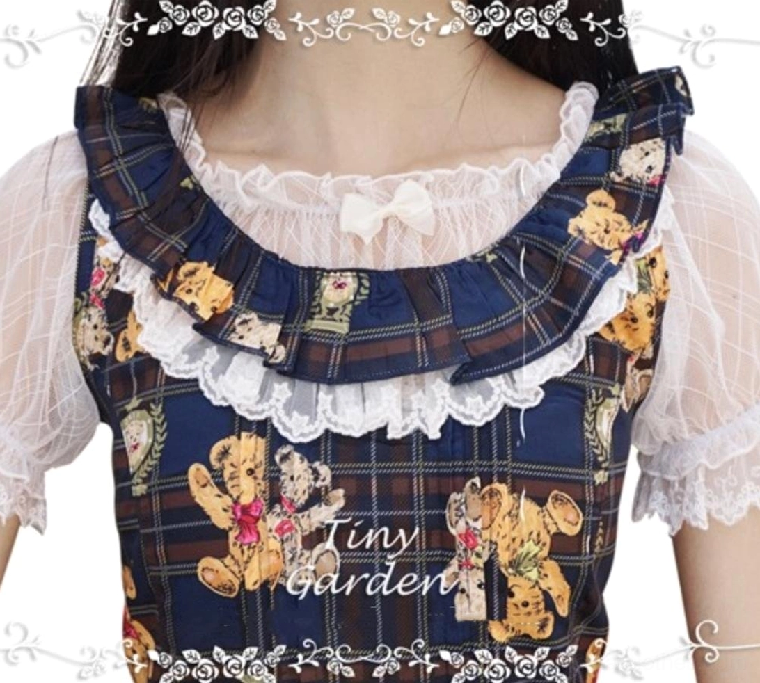 Model wearing white off-shoulder lolita inner blouse with ruffled neck bow and lace-trimmed short sheer sleeves, neck-down front close-up paired with teddy bear print plaid lolita jumpskirt