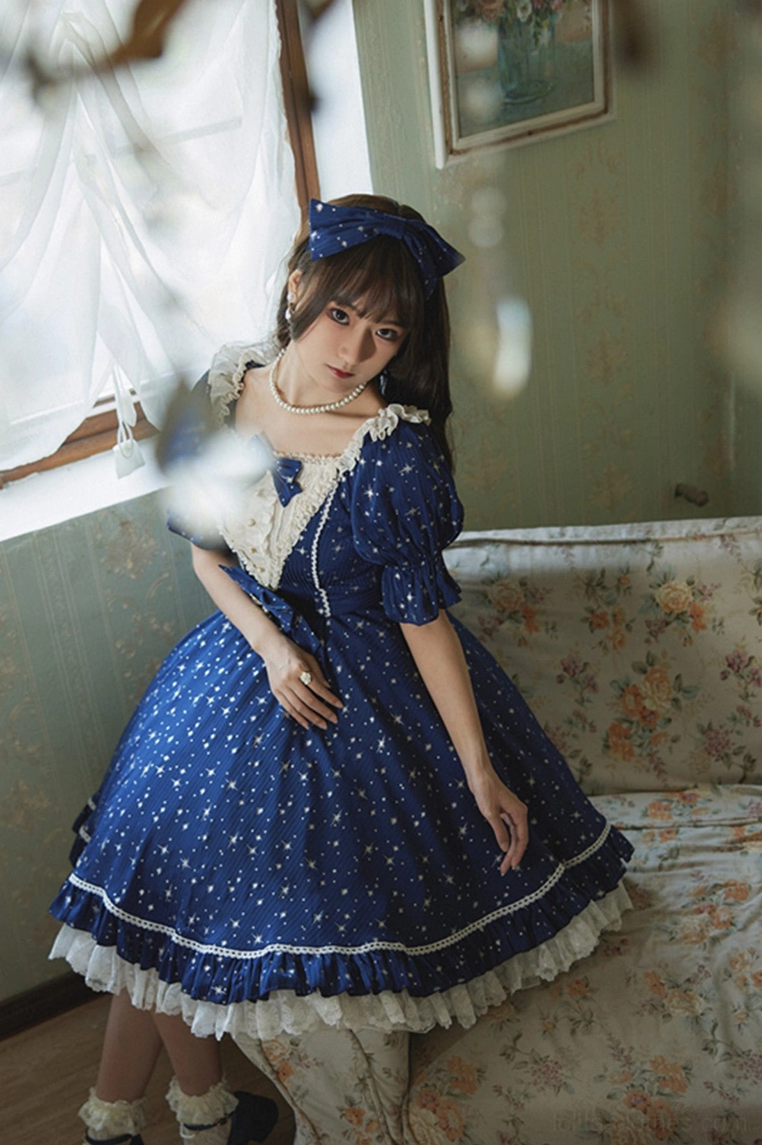 Navy blue star foil printed lolita jumper skirt dress half body sitting view vintage floral sofa matching hair bow white lace neck trim ruffled hem