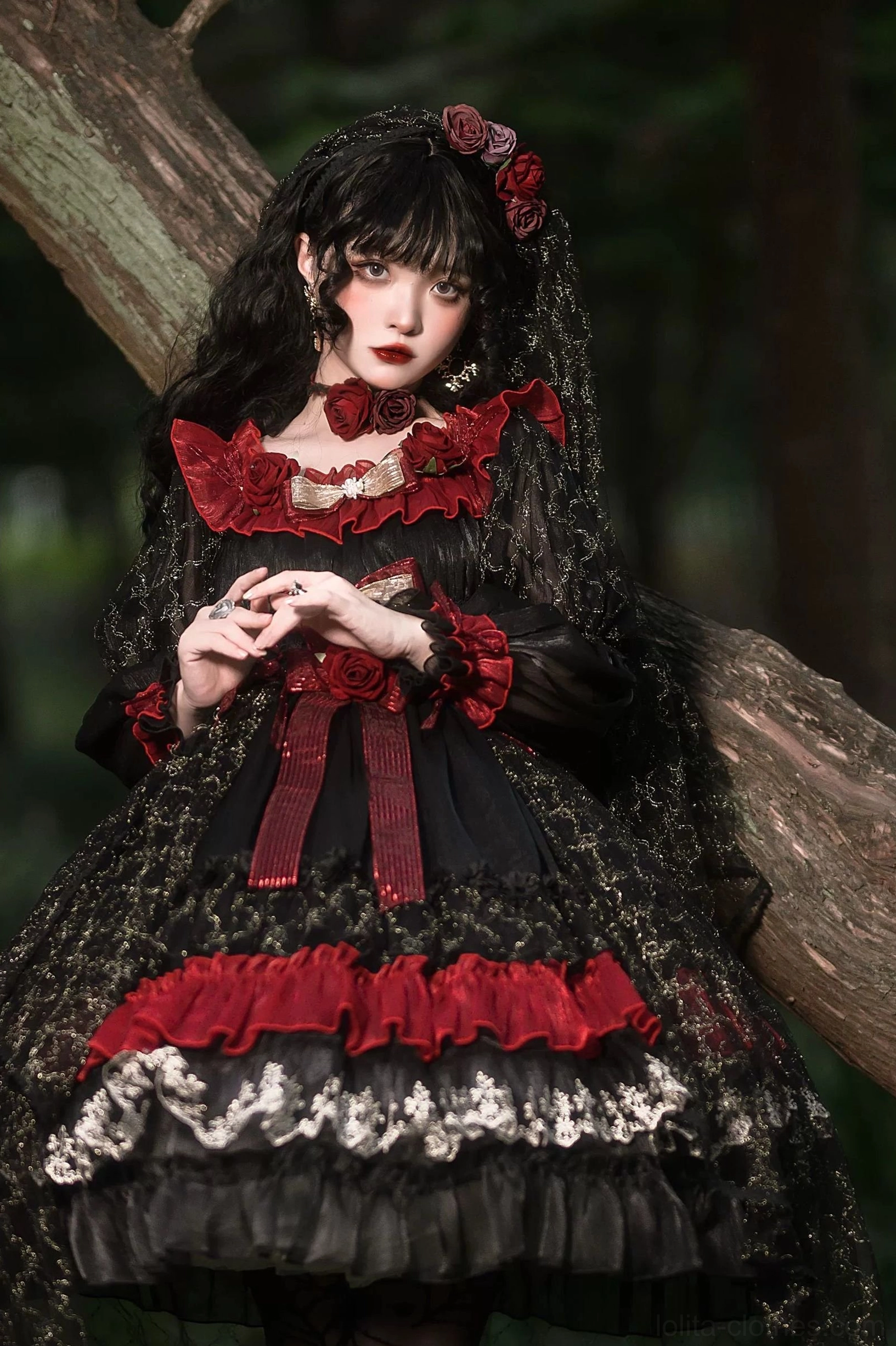 Gothic lolita OP dress seated half-body view in outdoor forest with tree backdrop, showing full layered skirt with red ruffles, silver lace trim, and matching rose hair veil