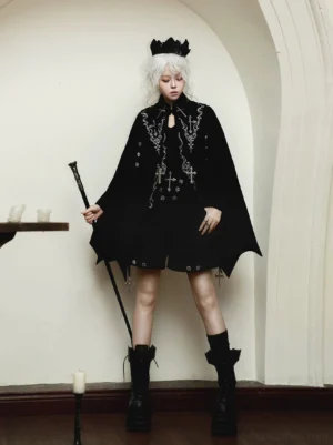 Full-body front view of gothic ouji lolita bat-wing cape outfit worn by white-haired model in black crown, featuring intricate silver cross embroidery on all-black ensemble with architectural cape and platform combat boots