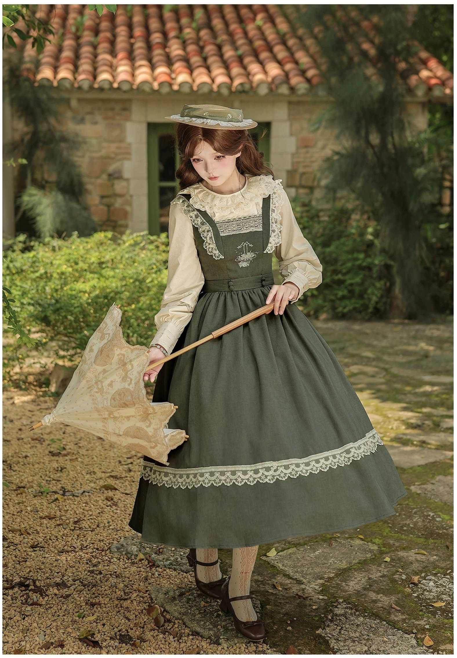 Single model wearing sage green lolita JSK with coordinating white lace blouse, front side full-body view in cottage courtyard with red tiled roof, holding vintage parasol accessory