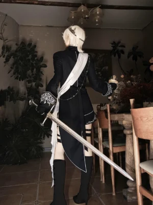 Oathbound noir ouji lolita military set back view showing dramatic high-low asymmetric tailcoat hemline, silver embroidery details, leg strap harness, black boots, vintage indoor furniture scene