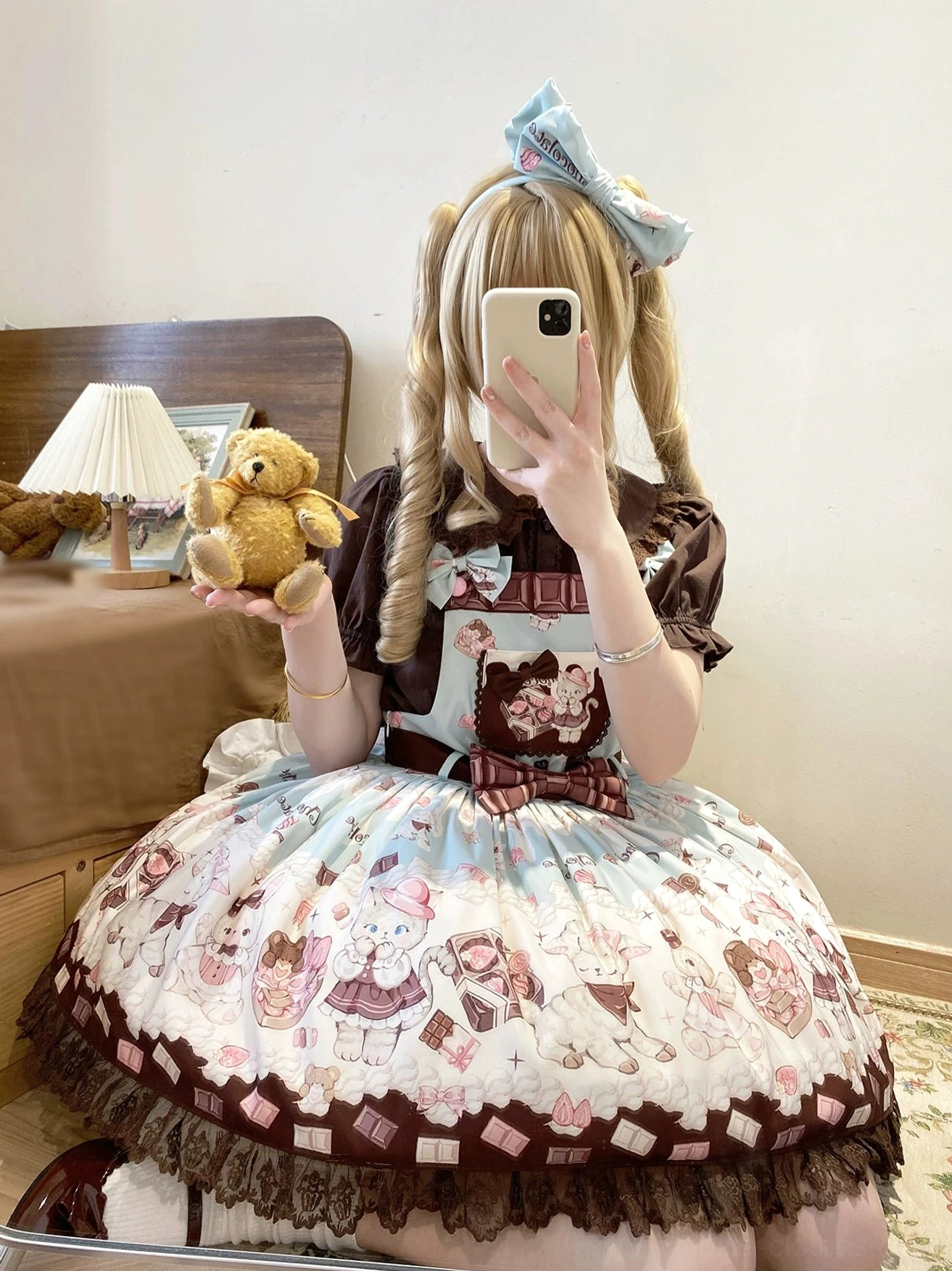 Blonde twin-tailed model taking mirror selfie wearing off-white and brown kawaii cat print Lolita salopette with blue hair bow, lace hemmed pleated skirt, and plush toy accessory