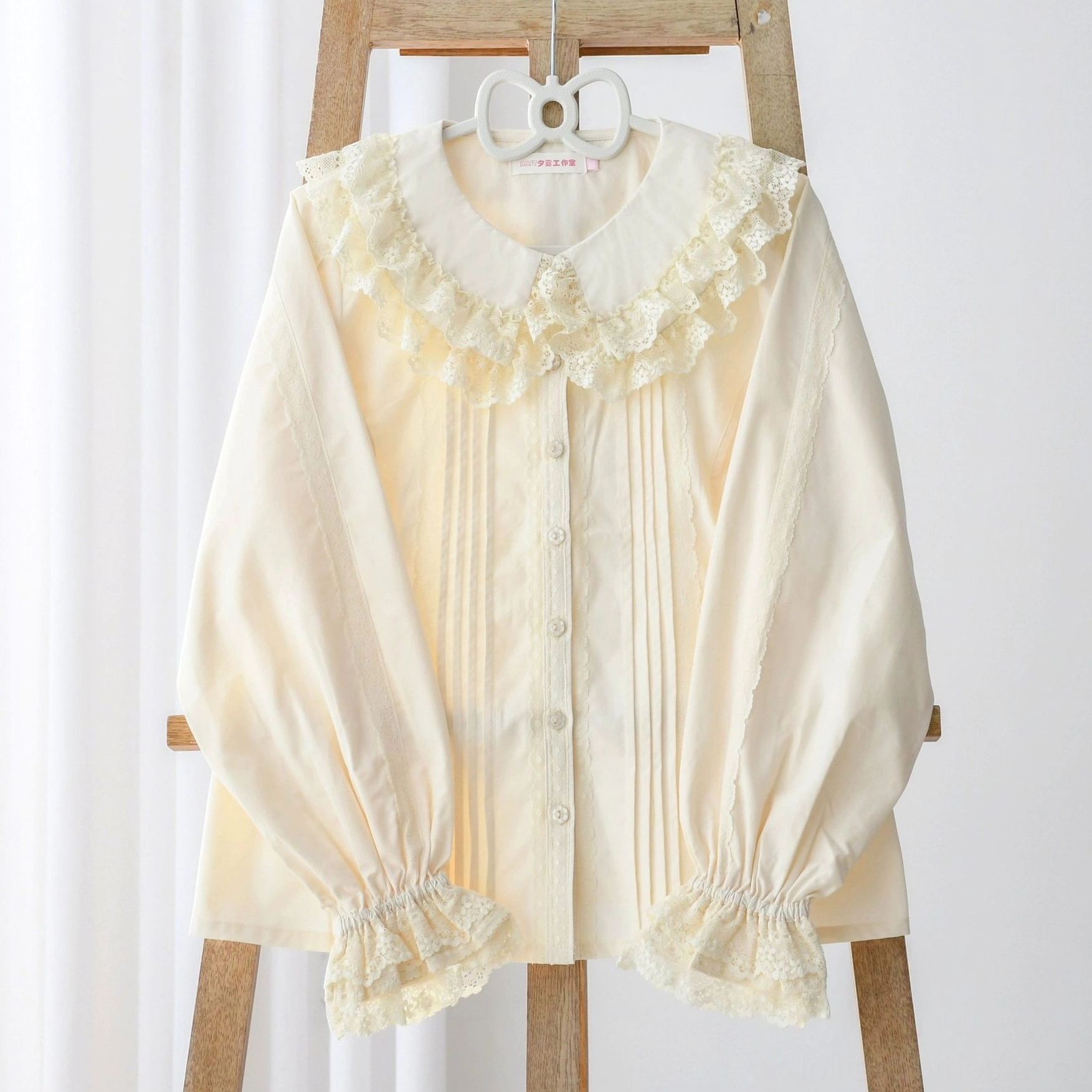 Close-up front flat display of off-white Lolita long-sleeve blouse on wooden hanger with white background, featuring lace ruffled collar and cuffs, front pleated button placket, and elegant lantern sleeves