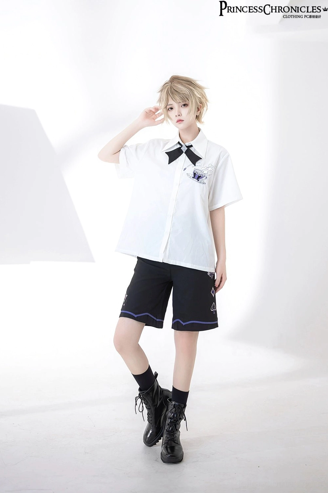 Short-haired androgynous model full body standing view wearing ouji embroidered short-sleeve shirt with black coordinating shorts and ankle boots