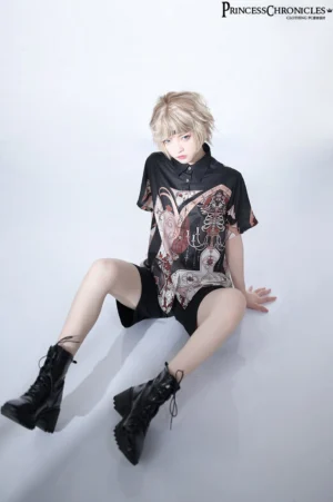 Ash blonde model wearing complete gothic Ouji Lolita coordinate featuring black printed shirt with crosses and vintage architecture designs paired with black shorts and coordinated retro floral hairband