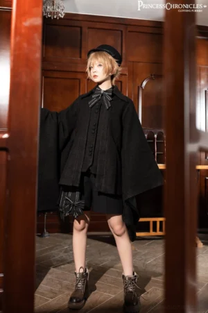 Full-body front view of aristocratic black winter cape coat ouji lolita worn by short-haired model in vintage wooden interior, featuring asymmetrical hemline, striped bow tie, wool beret, wide cape sleeves, decorative hem bow, paired with brown leather combat boots